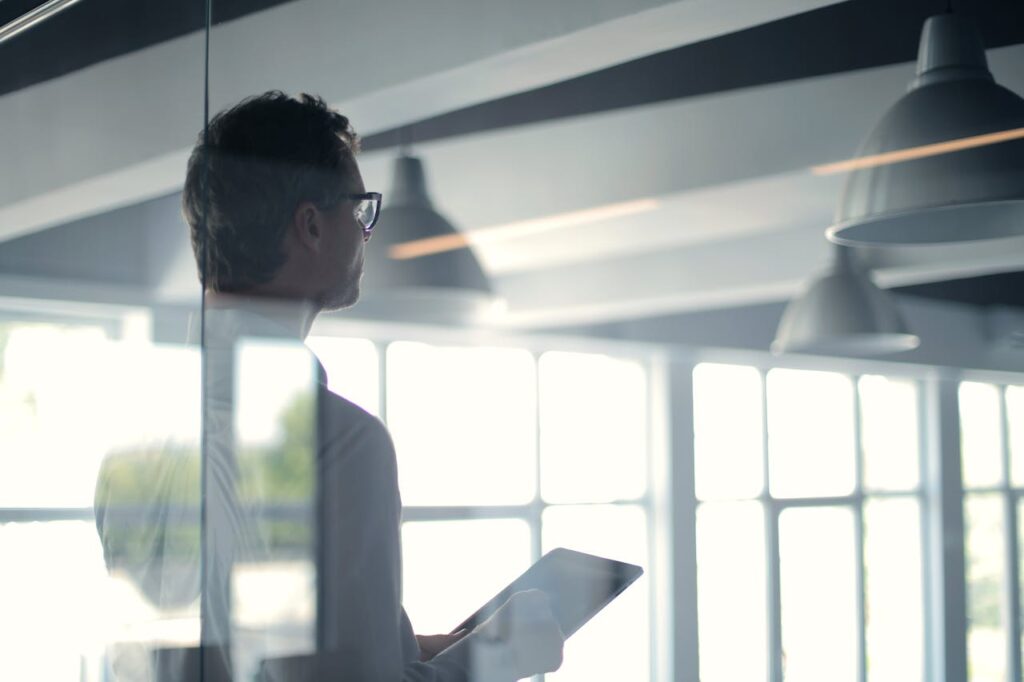 pexels photo 3760093 A businessman holding a tablet in an office, looking thoughtfully through a glass wall.